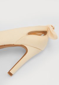 Scarpa con tacco beige caratterizzata da una finitura liscia, aperture laterali, un piccolo fiocco decorativo sul tallone e un design a stiletto affusolato.