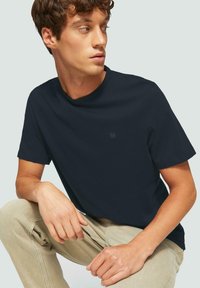 T-shirt a girocollo di colore blu navy in un tessuto morbido, maniche corte, con un logo discreto sul petto. Indossato con pantaloni beige chiaro.