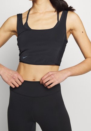 Femme portant un crop top de sport noir et des leggings taille haute, les mains posées sur les hanches, sur un fond clair uni.