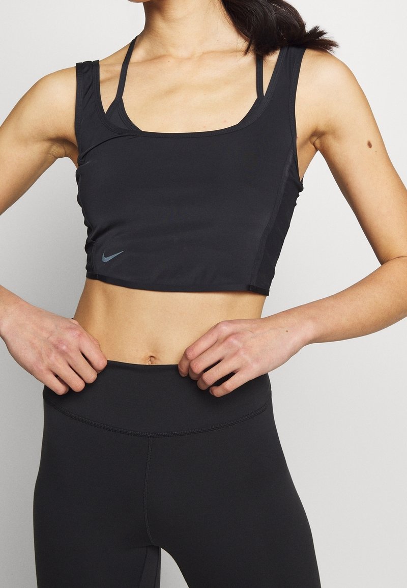 Femme portant un crop top de sport noir et des leggings taille haute, les mains posées sur les hanches, sur un fond clair uni.