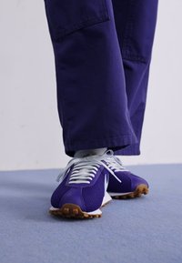 Paarse sportschoenen met witte veters en een witte swoosh, met een gestructureerde mesh bovenkant en een rubbere zool met bruine accenten.