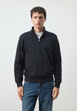 Homme aux cheveux courts et foncés portant un pull noir à demi-fermeture éclair sur une chemise blanche et un jean bleu, debout devant un fond uni.