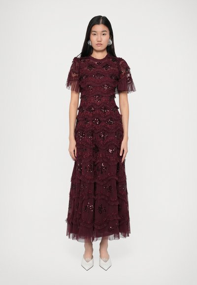 Femme portant une longue robe en dentelle bourgogne avec des volants et des sequins, associée à des talons blancs pointus et des boucles d'oreilles en argent.
