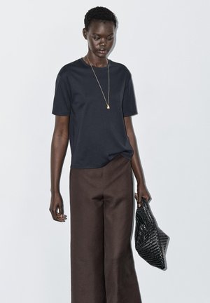 Woman wearing a black short-sleeve top and brown wide-leg pants, holding a black woven clutch bag, against a light gray background.