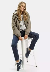 Khaki kurtka z kapturem, szara bluza pod spodem, ciemne dżinsy i granatowe sneakersy z białymi akcentami, siedzący na białym stołku.