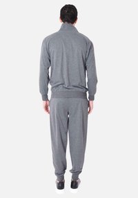 Tuta grigia con colletto alto, polsini a coste e vita elasticizzata, realizzata in un tessuto morbido e liscio. Indossata con scarpe nere.