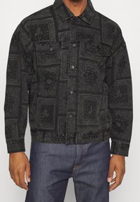 Veste en denim noir avec une texture à motifs, dotée d'un col classique, d'une fermeture à boutons et de poches sur la poitrine. Agrémentée de dessins en relief.