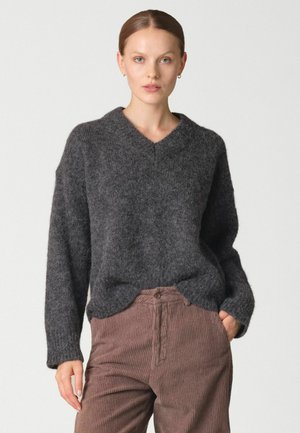 Femme portant un pull à col en V duveteux gris foncé et un pantalon en velours côtelé marron, debout avec une main dans la poche sur un fond clair.