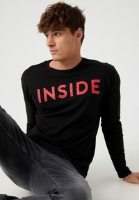 Camiseta negra de manga larga con un bold texto rojo "INSIDE", combinada con unos vaqueros negros desgastados, que presentan un corte ajustado y una textura de tela suave.