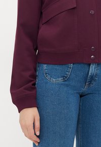 Maroon cotton jacket with front pockets, snap button closures, and ribbed cuffs, paired with blue denim jeans featuring back pockets.
