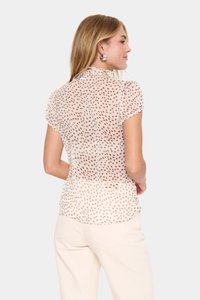 Durchsichtige beigefarbene Bluse mit kurzen Ärmeln, die ein gepunktetes Muster in Braun aufweist. Weicher Stoff mit Rüschenausschnitt und leicht taillierter Silhouette.