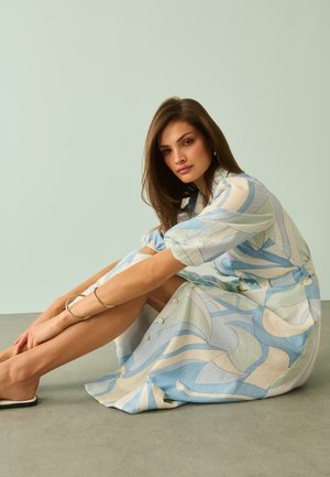 Fiatal nő hosszú barna hajjal, világoskék és krémszínű mintás ruhát visel, lapos, belebújós cipőkkel, miközben a padlón ül.