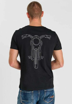 Man met de rug naar voren, draagt een zwart T-shirt met een witte contour van het vooraanzicht van een motorfiets op de achterkant, gecombineerd met blauwe spijkerbroek.