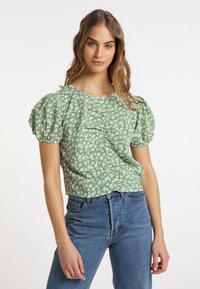 Grünes floral gemustertes Kurzarmoberteil mit geschöpften Ärmeln, das eine strukturierte Oberfläche und einen runden Halsausschnitt aufweist, kombiniert mit blauen hochgeschnittenen Jeans.
