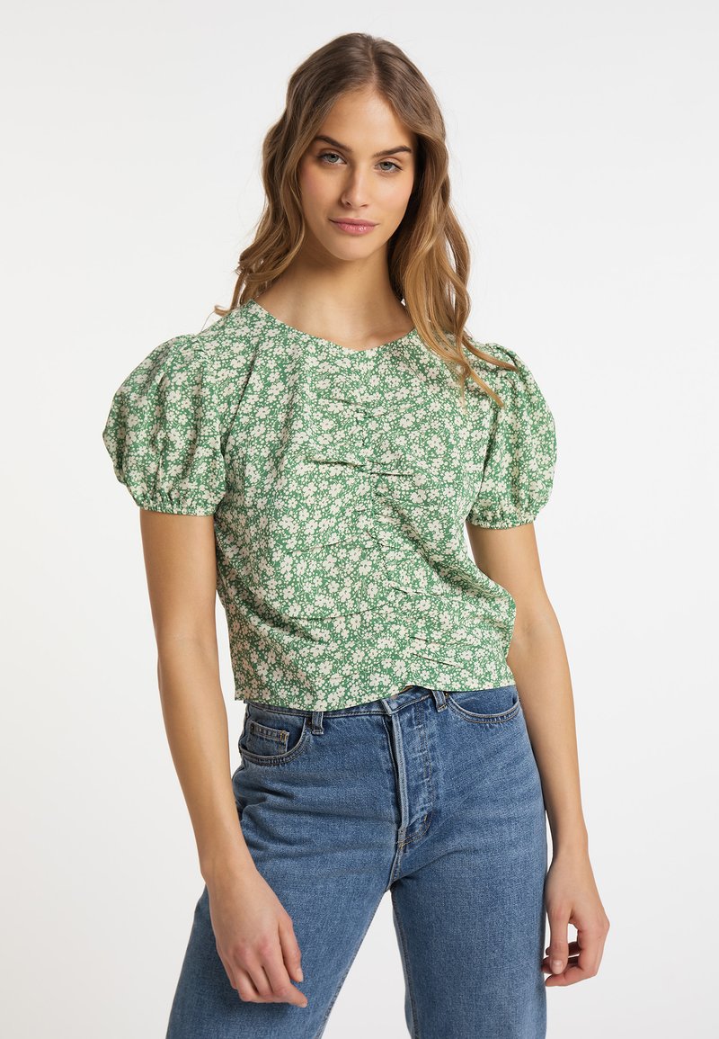 Grünes floral gemustertes Kurzarmoberteil mit geschöpften Ärmeln, das eine strukturierte Oberfläche und einen runden Halsausschnitt aufweist, kombiniert mit blauen hochgeschnittenen Jeans.