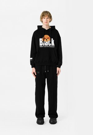 Sudadera negra con estampado gráfico y bolsillo canguro, combinada con pantalones de chándal negros con bolsillos con cremallera. Llevado con zapatillas negras.