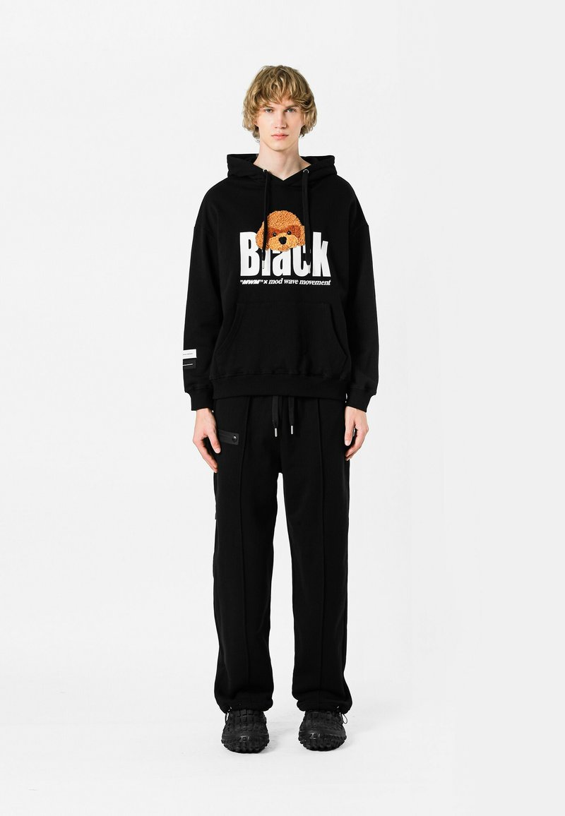 Sudadera negra con estampado gráfico y bolsillo canguro, combinada con pantalones de chándal negros con bolsillos con cremallera. Llevado con zapatillas negras.