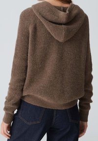 Osoba w brązowej, puchatej bluzie z długimi rękawami i ciemnoniebieskich dżinsach, pokazana z tyłu na jednolitym tle.