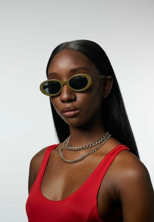 Olijfgroene zonnebril met een ovale montuur, donkere lenzen en metalen accenten. Ggedragen met een rode mouwloze top en een gelaagde zilveren ketting.