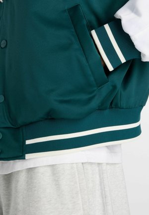 Mørkegrøn varsityjakke med hvide stribede ribkanter ved ærmer og kant, hånd i sidelomme, båret over en hvid skjorte og lysegrå bukser.