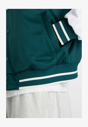 Mørkegrøn varsityjakke med hvide stribede ribkanter ved ærmer og kant, hånd i sidelomme, båret over en hvid skjorte og lysegrå bukser.