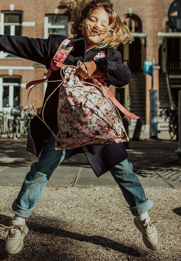 Rugzak met een pink, patroonstof met speelse dieren; zacht, ruim ontwerp met roze banden; kind dat vrolijk buiten aan het springen is.