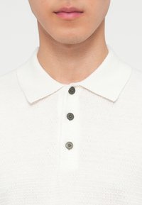 Homme portant un polo en tricot blanc avec trois boutons noirs, photographié du menton à la poitrine.