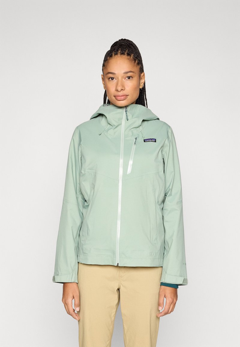 Casaco impermeável verde claro com capuz, apresentando um fecho éclair frontal, bolsos laterais e um logotipo no peito. Textura suave e design prático.