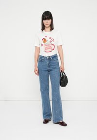 Kvinne iført en hvit høytids-tema T-skjorte med design av en pepperkakekrus, blå jeans, burgunder sko, og holder en svart veske.