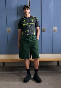 Camiseta deportiva negra y verde neón, pantalones cortos verdes, zapatos negros sin cordones, de pie frente a taquillas azules. Banco de metal simple debajo.