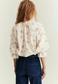 Hvid blomstret bluse med flagrende ærmer, samlet halsudskæring og tekstureret stof, parret med mørk denim jeans med forlommer.