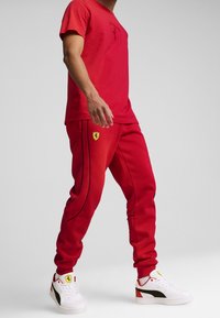 Man med röd Ferrari-tryckt t-shirt och joggers med vita Puma-sneakers som har Ferrari-logotyper mot en enkel grå bakgrund.