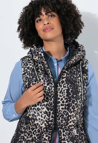 Weste mit Leopardenmuster, ausgestattet mit einer Kapuze, schwarzem Reißverschluss und gebrandeten Riemen. Über einem blau-weiß gestreiften Hemd getragen. Glatte, gesteppten Textur.