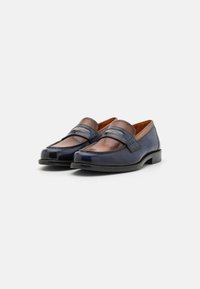 Mocassins en cuir bleu et marron avec une texture lisse, des coutures contrastantes et une bande décorative sur le devant. Bout rond et petit talon.
