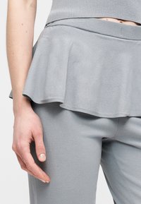 Γκρι τοπ πεπλούμ με ριμπ υφή, στρωτή γαζιά και εφαρμοστά γκρι παντελόνια. Το απαλό ύφασμα αντανακλά διακριτικό φως, αποπνέοντας απαλή εμφάνιση.