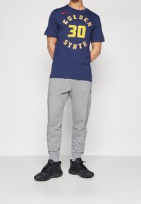 Camiseta navy con "GOLDEN STATE" y "30" en amarillo y dorado, combinada con pantalones de chándal gris ajustados y zapatillas deportivas negras.