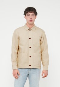 Μπεζ τζιν μπουφάν με καφέ κουμπιά, γιακά και δύο μπροστινές τσέπες. Διαθέτει ραφές στο επάνω μέρος και μια μικρή ετικέτα με επωνυμία.