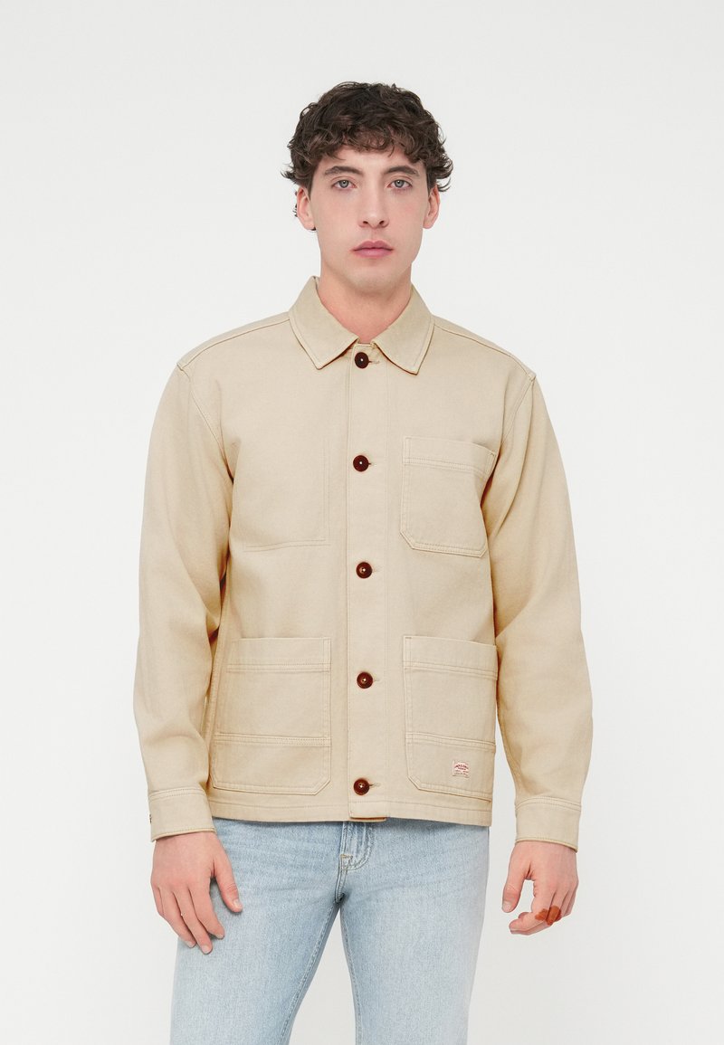 Μπεζ τζιν μπουφάν με καφέ κουμπιά, γιακά και δύο μπροστινές τσέπες. Διαθέτει ραφές στο επάνω μέρος και μια μικρή ετικέτα με επωνυμία.