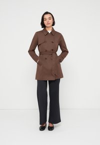 Καφέ διπλόμπουφο trench coat με ζώνη, μεγάλο γιακά και κουμπιά ως διακοσμητικά στοιχεία, συνδυασμένο με μαύρο παντελόνι με ρίγες και μαύρα παπούτσια.