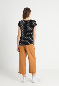 Svart topp med vit diagonalrand, korta ärmar och knapppå detalj på baksidan, i kombination med lösa bruna byxor och vita sneakers.