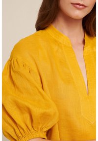 Blusa di lino gialla con maniche a sbuffo, scollatura a V e finitura texturizzata. Presenta dettagli cuciti lungo il colletto e le maniche.