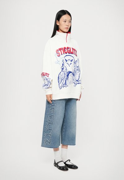 Felpa bianca con grafica di un'aquila blu e testo rosso "STIGLITZ", abbinata a pantaloni di denim larghi e cropped con testo blu. Scarpe nere e calze bianche.