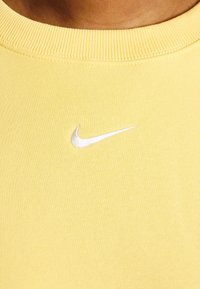 Gul bomullströja med ribbad krage och en liten vit Nike-logotyp broderad på framsidan. Slät textur, avslappnad design.