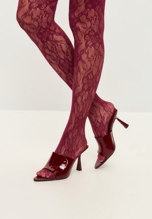 Piernas con medias de encaje floral burdeos que llevan mules de tacón abiertos y brillantes, cruzadas en los tobillos sobre un fondo blanco.