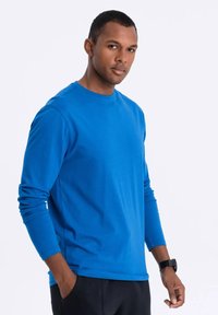 Pitkähihainen, pyöreäkauluksinen t-paita kirkkaan sinisestä kankaasta, joka on sileä ja rennosti istuva. Yhdistetty mustiin housuihin ja kelloon.