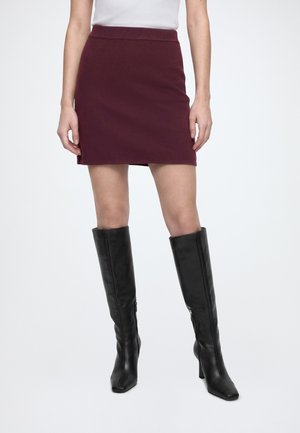 Femme portant une mini-jupe bordeaux, un haut blanc et des bottes noires à talons hautes jusqu'aux genoux, debout devant un fond uni.