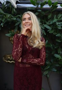 Vinrdeča čipkasta obleka z dolgimi rokavi in cvetličnimi vzorci, z oprijeto silhueto in prozorno plastjo, postavljena na zeleno listje.