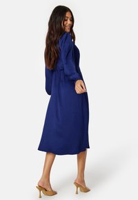 Středně dlouhé, tmavě modré šaty s dlouhými nafouklými rukávy, upnutým pasem a texturovanou tkaninou, doplněné béžovými sandály na vysokém podpatku.