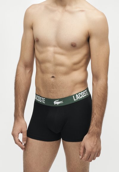 Мъж без риза, носещ черни боксерки Lacoste със зелен колан и бяло лого, стои пред обикновен светъл фон.