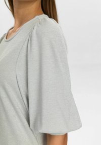 Femme portant un haut gris clair en tissu côtelé avec des manches courtes bouffantes, montrant le buste et le bras contre un fond uni.